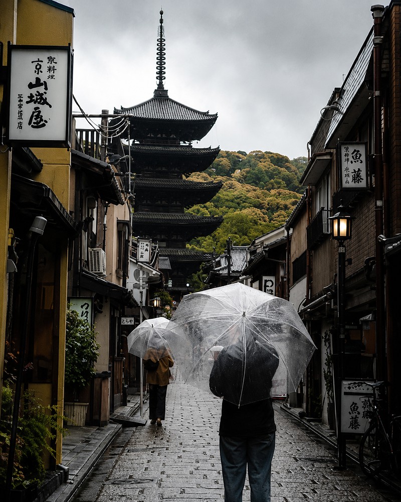 Pessoas caminhando sob guarda-chuvas transparentes em uma rua tradicional de Quioto, em dia de chuva, com um pagode ao fundo
