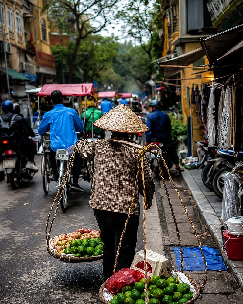 Vendedora ambulante carregando cestos de frutas por uma rua movimentada em Hanói